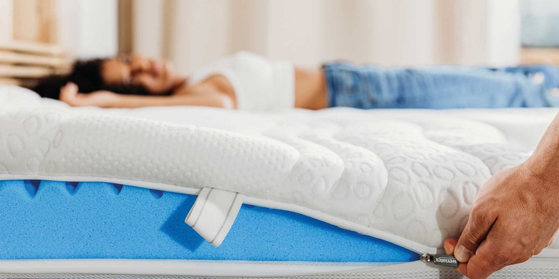 Mann öffnet den Bezug einer Wenatex Matratze Comfort Plus, während eine Frau im Hintergrund entspannt liegt.