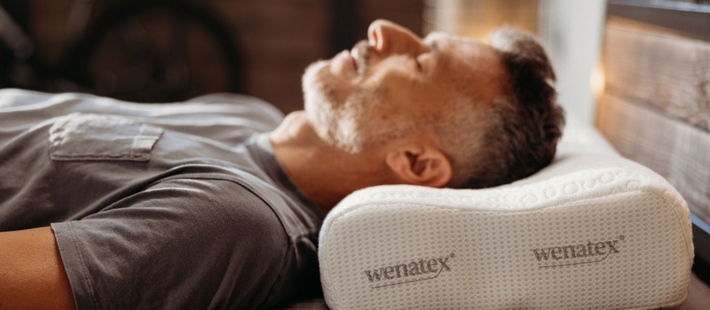 Mann liegt entspannt mit dem Wenatex Nackenkissen Wenacel Comfort auf einem Bett.